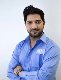 Ein Mann mit dunkelhaariger Frisur steht mit verschränkten Armen in blauem Hemd vor weißer Wand.