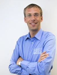 Mann mit Brille und blauem Hemd steht lächelnd mit verschränkten Armen vor weißer Wand.