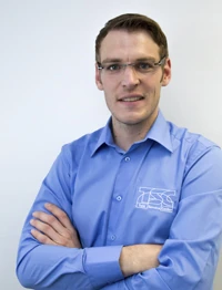 Mann mit Brille und blauem Hemd steht vor weißem Hintergrund und blickt freundlich.